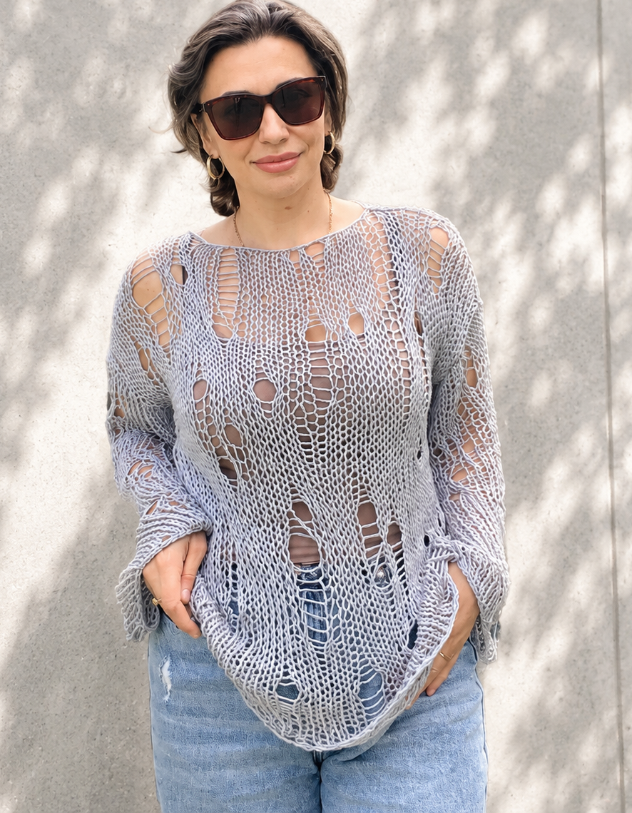 Woman wearing an oversized distressed cotton knit sweater in soft grey blue color with relaxed luxury styling against a light concrete wall with natural shadows