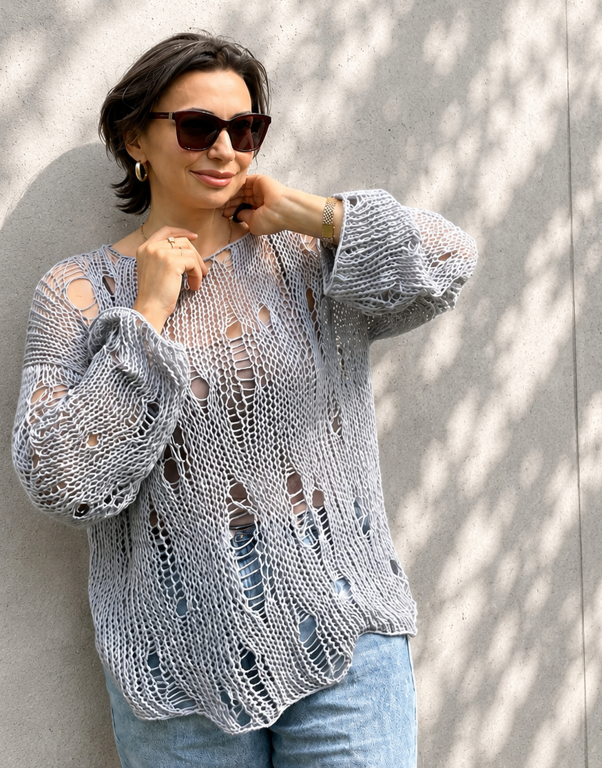 Woman wearing an oversized distressed cotton knit sweater in grey blue color with airy loose knit texture and relaxed luxury summer styling against a light concrete wall with soft natural shadows