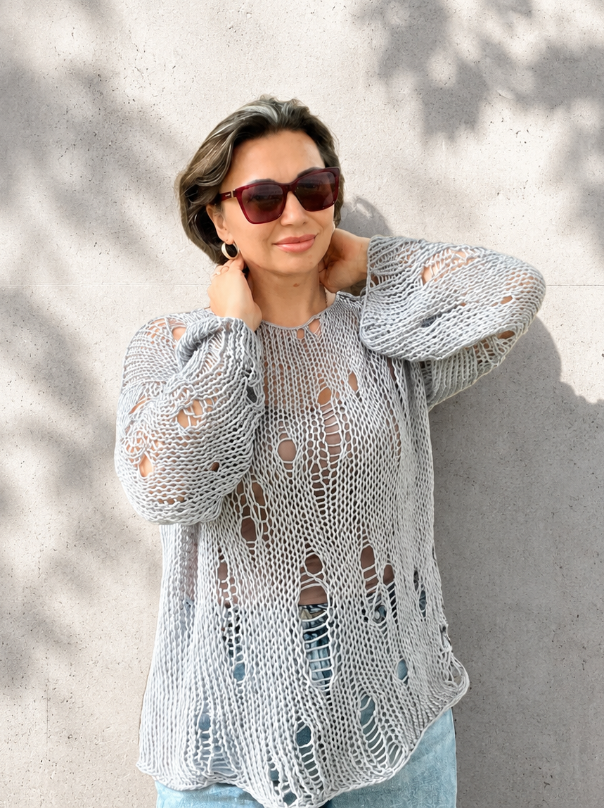 Woman wearing an oversized distressed cotton knit sweater in grey blue color with relaxed luxury summer styling against a light concrete wall with soft natural shadows