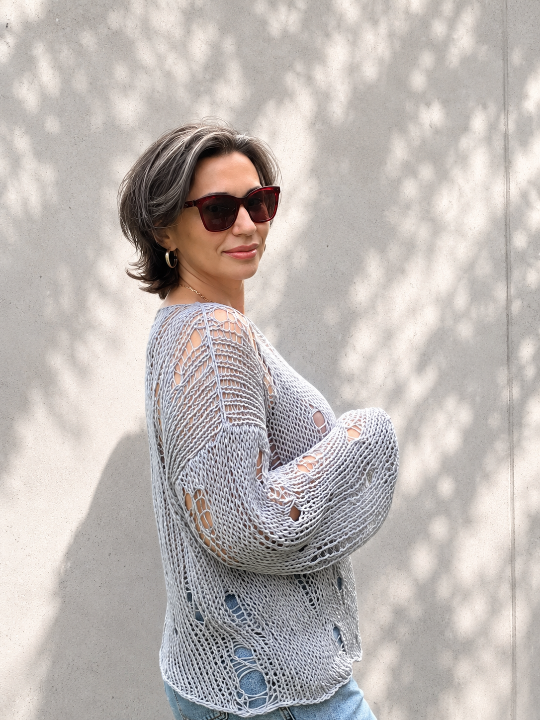 Close-up of an oversized distressed cotton knit sweater in grey blue color with airy open-knit texture and relaxed luxury summer styling against a light concrete wall with soft natural shadows
