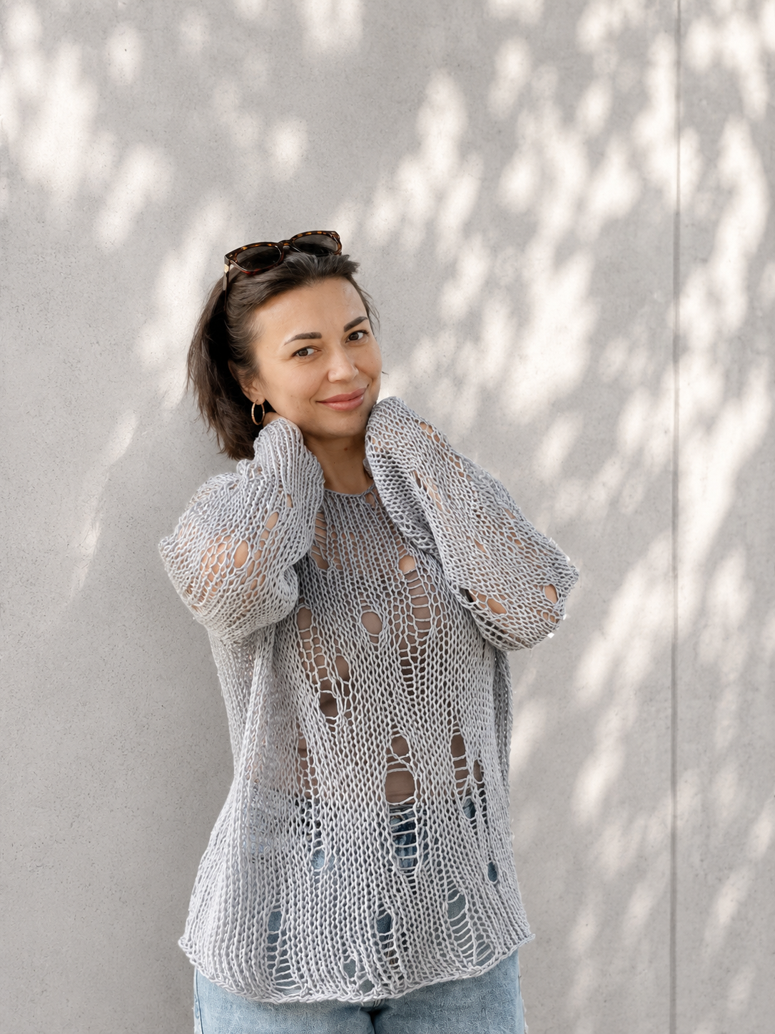 Woman wearing an oversized distressed cotton knit sweater in soft grey blue color with airy open-knit texture and relaxed luxury summer styling against a light concrete wall with natural shadows